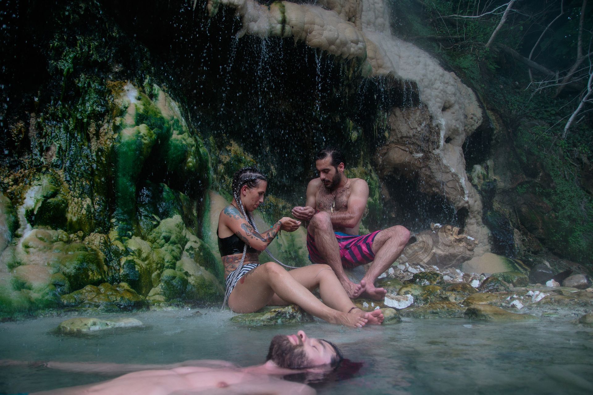 People sit and swim in a natural thermal spring surrounded by lush, mossy rock formations under a gentle waterfall.