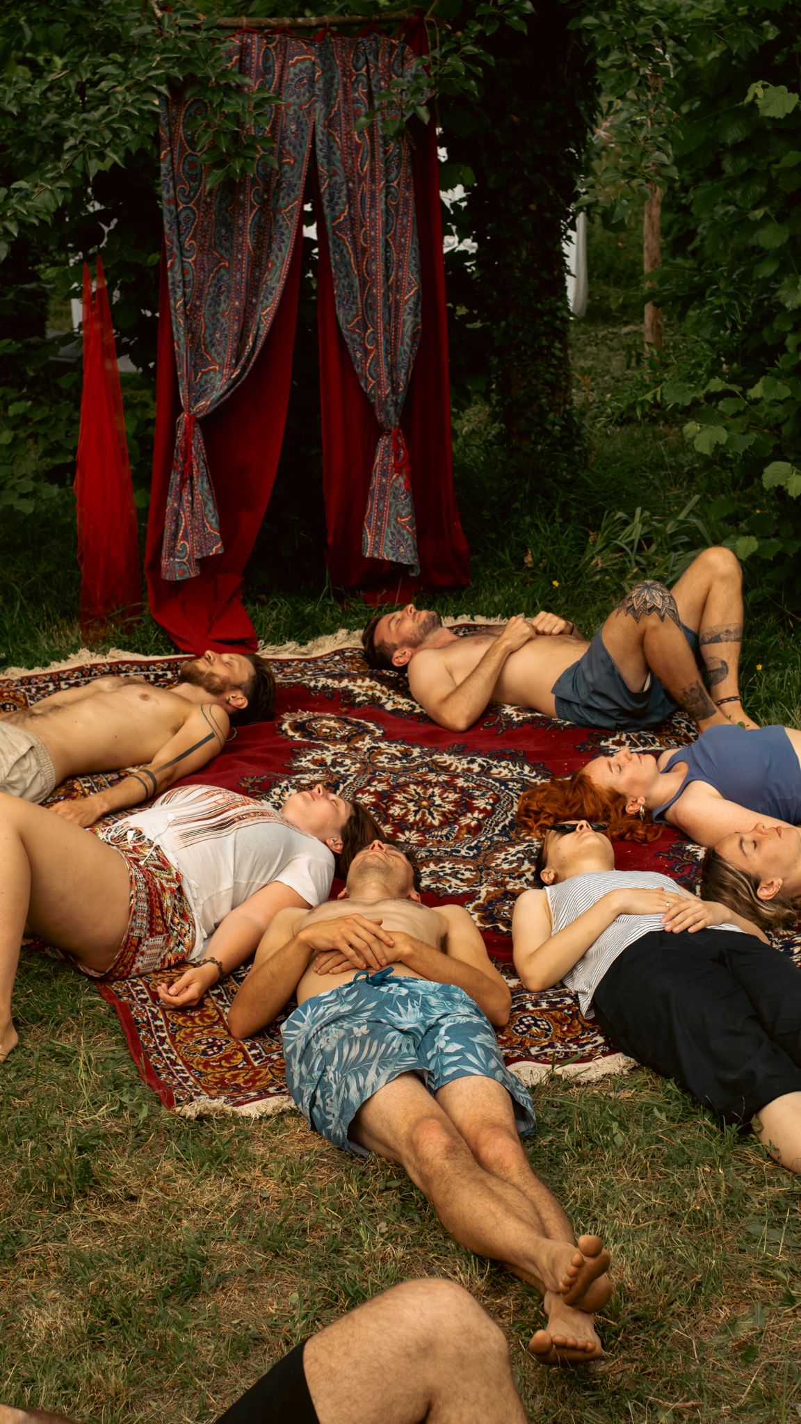 A group of people lying on rugs in a grassy outdoor area near a red and patterned fabric backdrop.