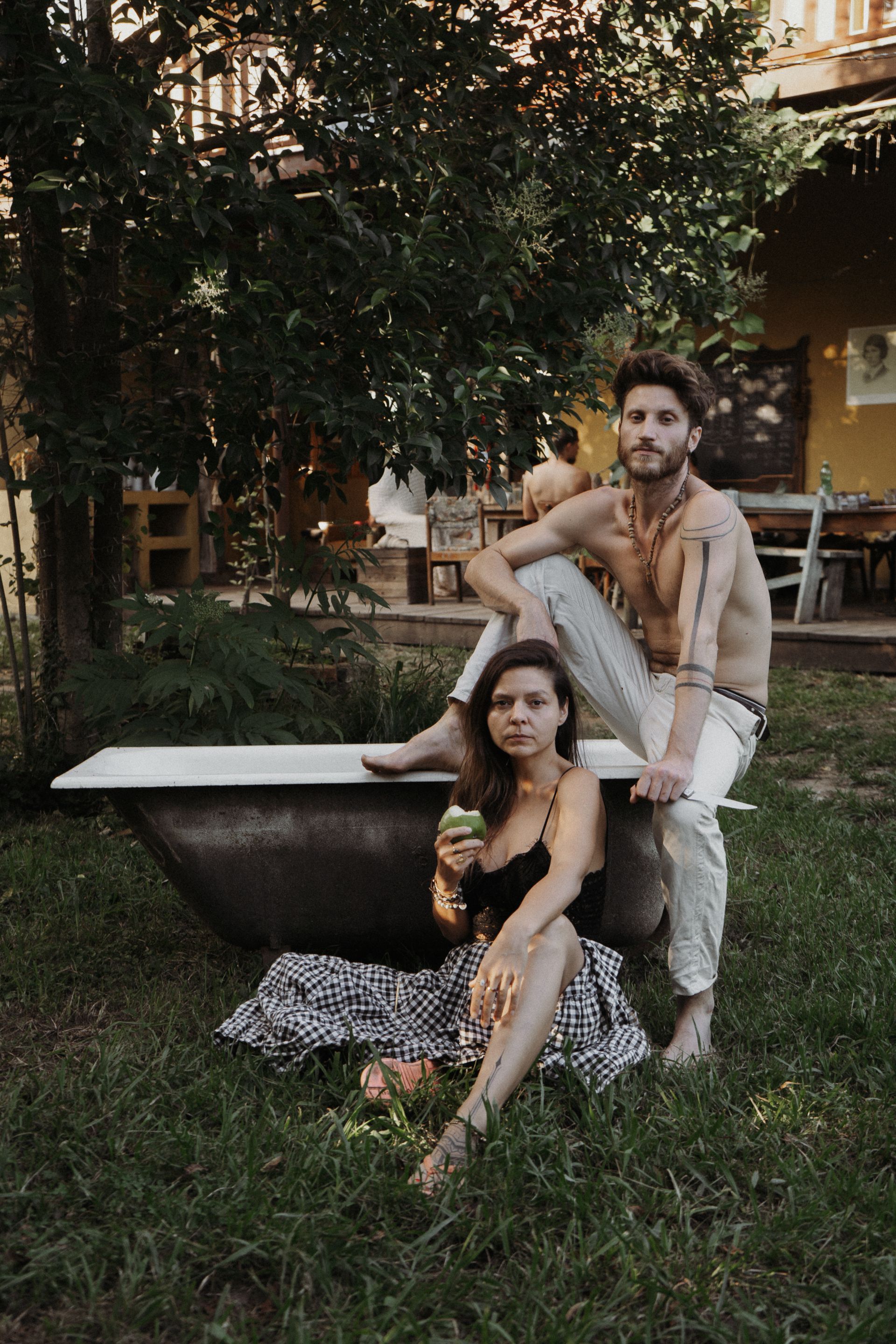 A person sits inside a rustic bathtub in a garden, while another sits on the edge holding a green drink.