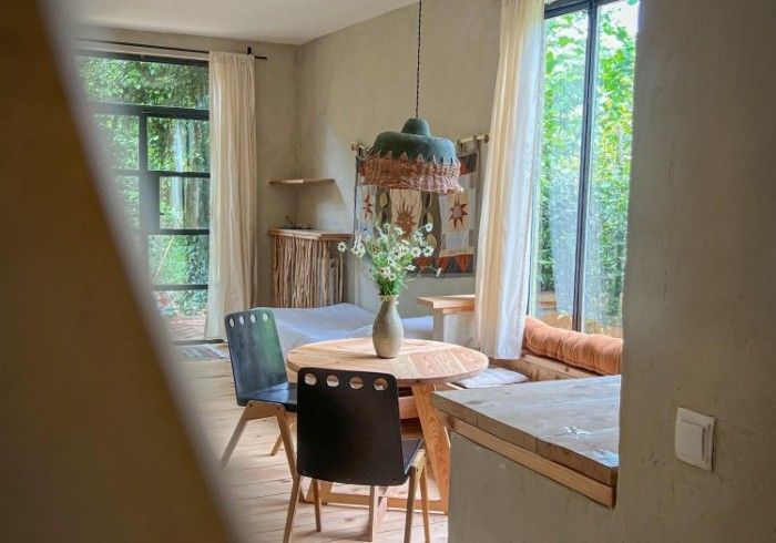A sunlit dining nook with a round wooden table, two chairs, a pendant lamp, and a bench by large windows looking outdoors.