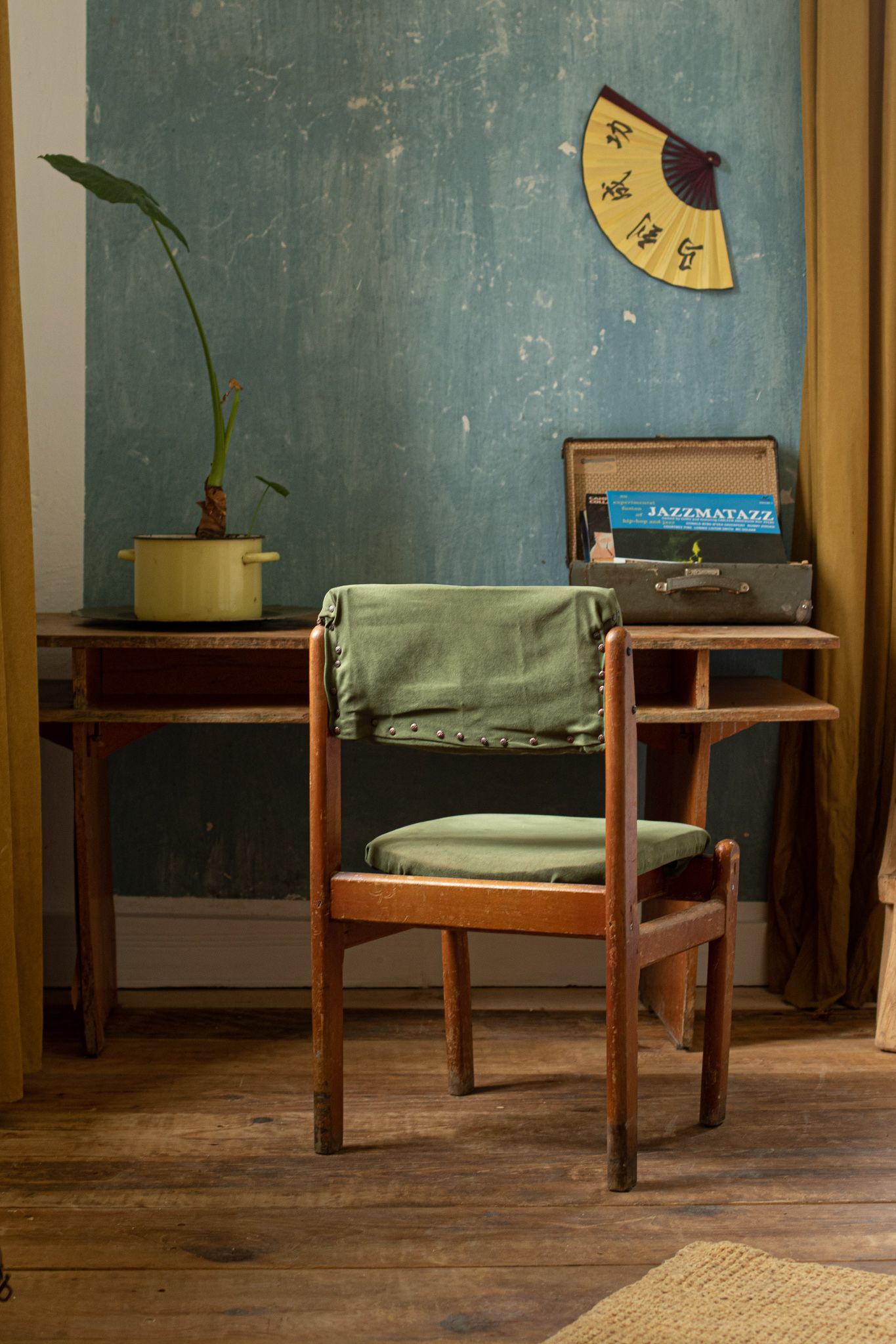 A rustic wooden desk with a chair, green cushions, a potted plant, and a vintage suitcase against a textured teal wall.