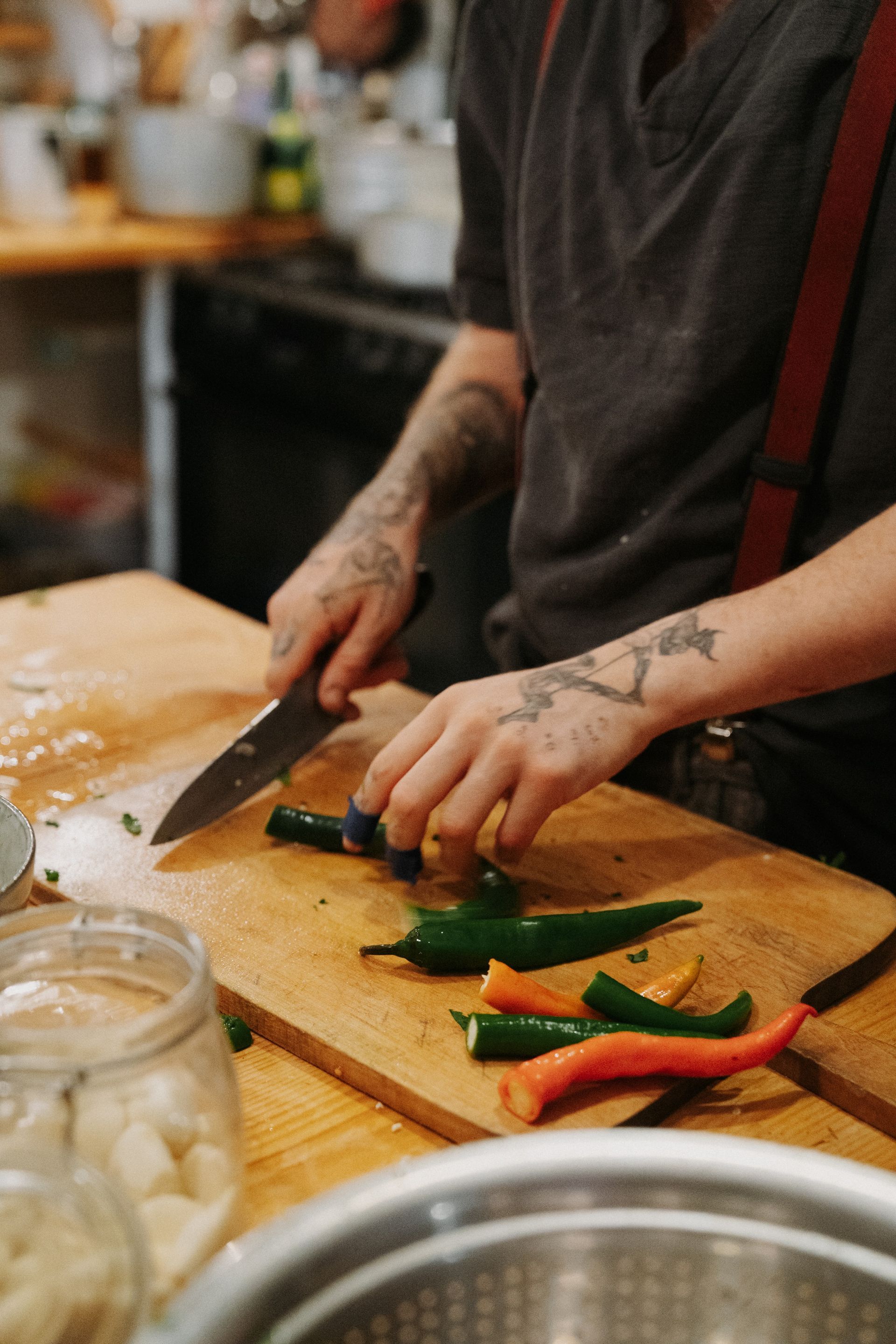 A person with tattoos on their hands and arms uses a knife to slice green peppers on a wooden cutting board in a kitchen.