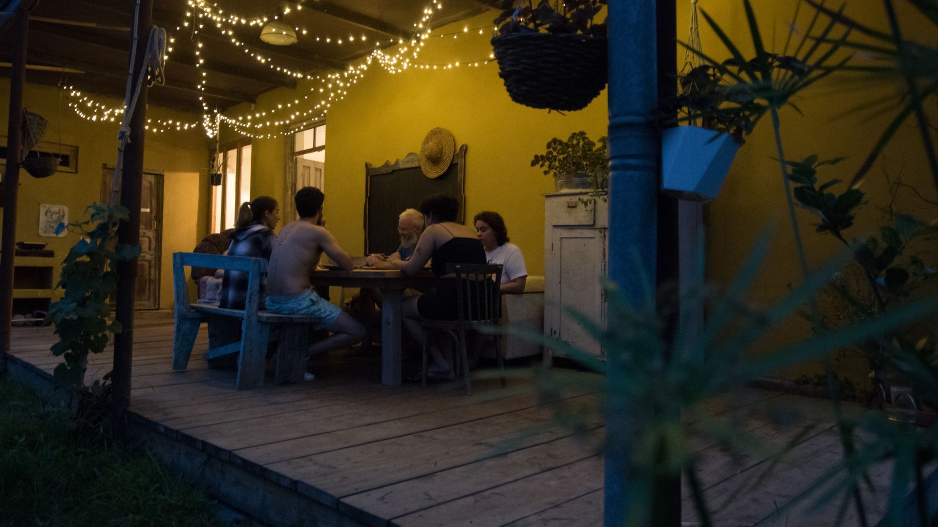 A group of people sit around a table on a yellow-walled outdoor patio decorated with fairy lights at dusk.