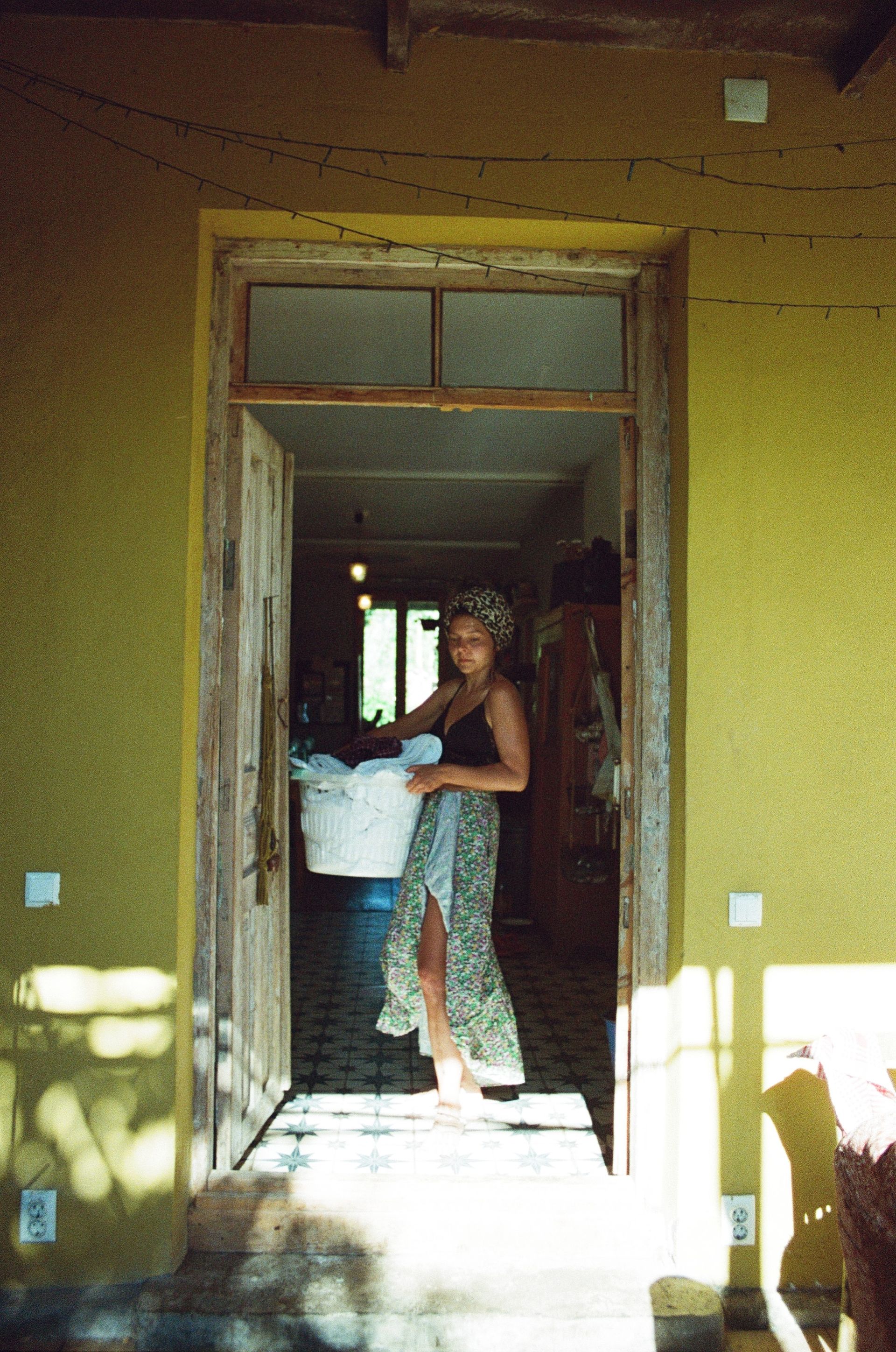 A person stands in a doorway holding a basket of laundry, wearing a black top and patterned skirt against a yellow wall.