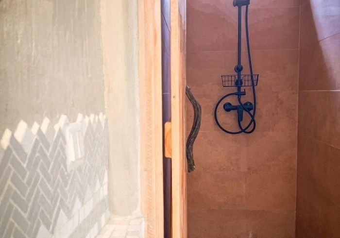 A shower stall with tan walls, a black shower fixture, and a wooden door frame, beside a wall with gray herringbone tiles.