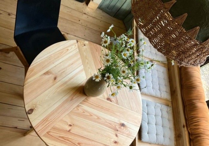 A top-down view of a round, light-wood table with a vase of daisies, set next to a chair and a cushioned wooden bench.