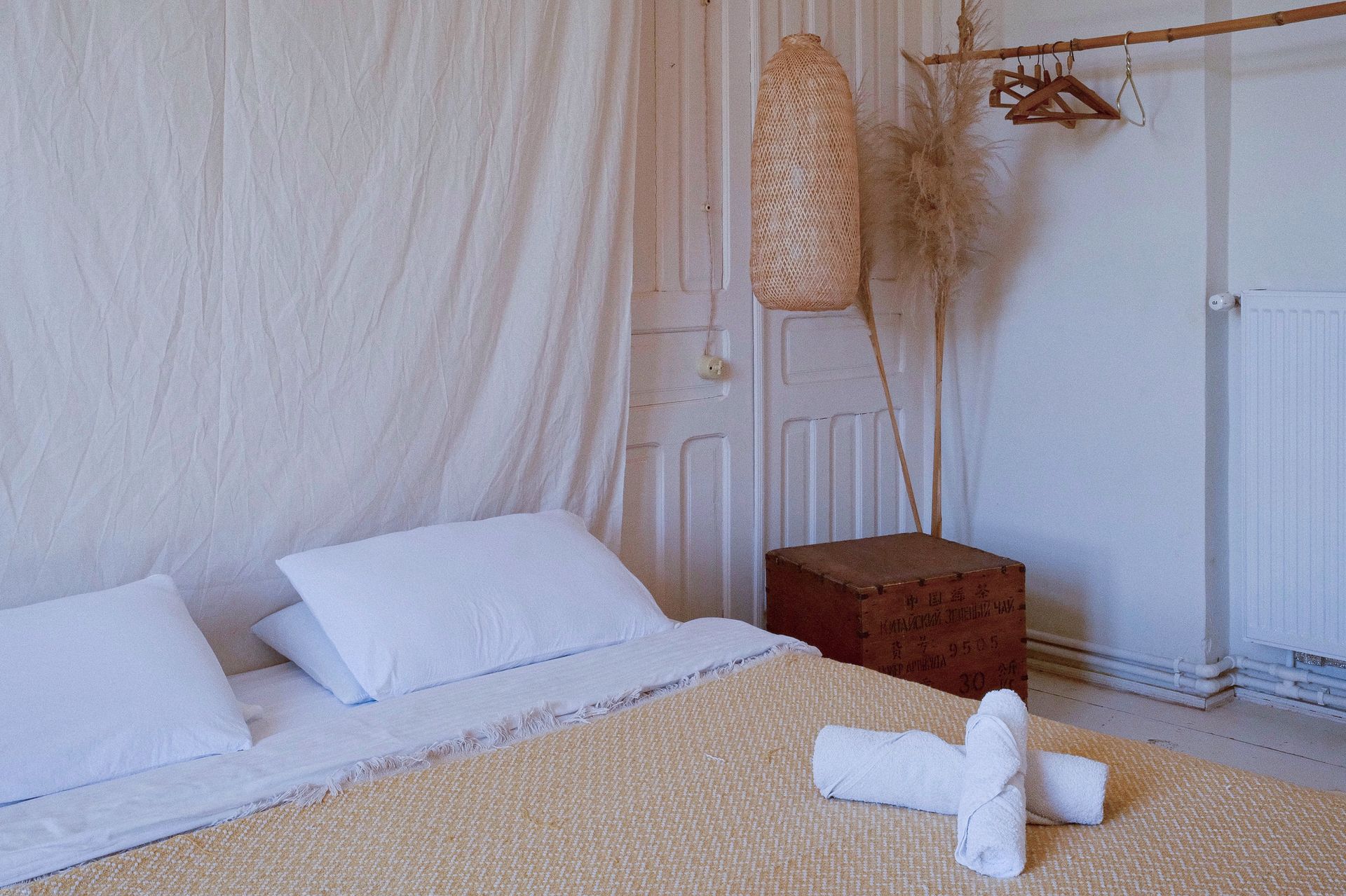 A minimalist bedroom with white walls, a textured bedspread, rolled white towels, a rattan light, and a wooden clothes rack.
