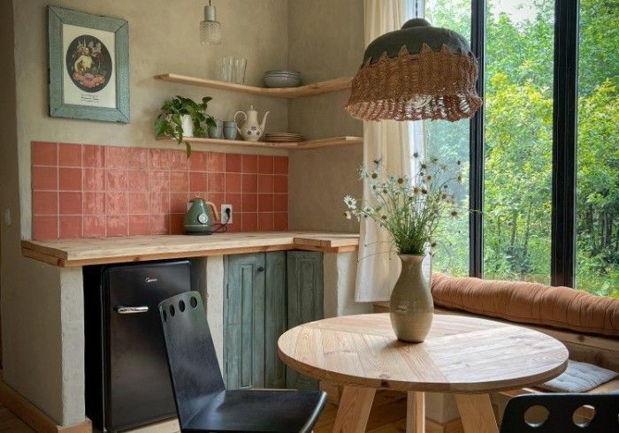 A rustic kitchen corner with a wooden table, black chairs, terracotta backsplash, open shelves, and a large window.