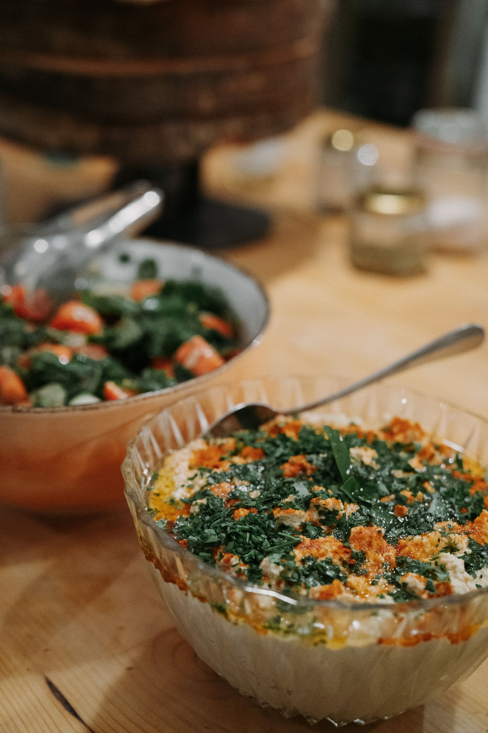 Two glass bowls on a wooden table, one filled with a tomato and herb salad, the other with a layered dip topped with greens.