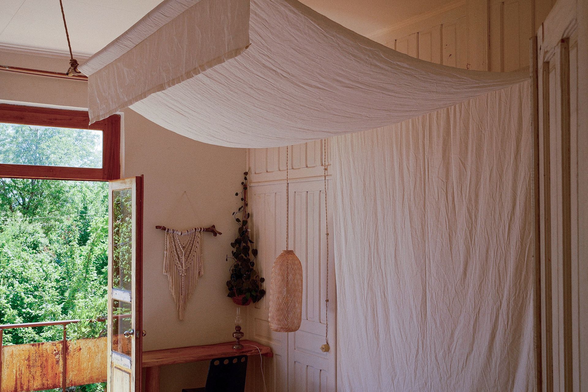 A canopy of draped white fabric hangs over a room corner with a balcony door, a wall hanging, and a small wooden desk.