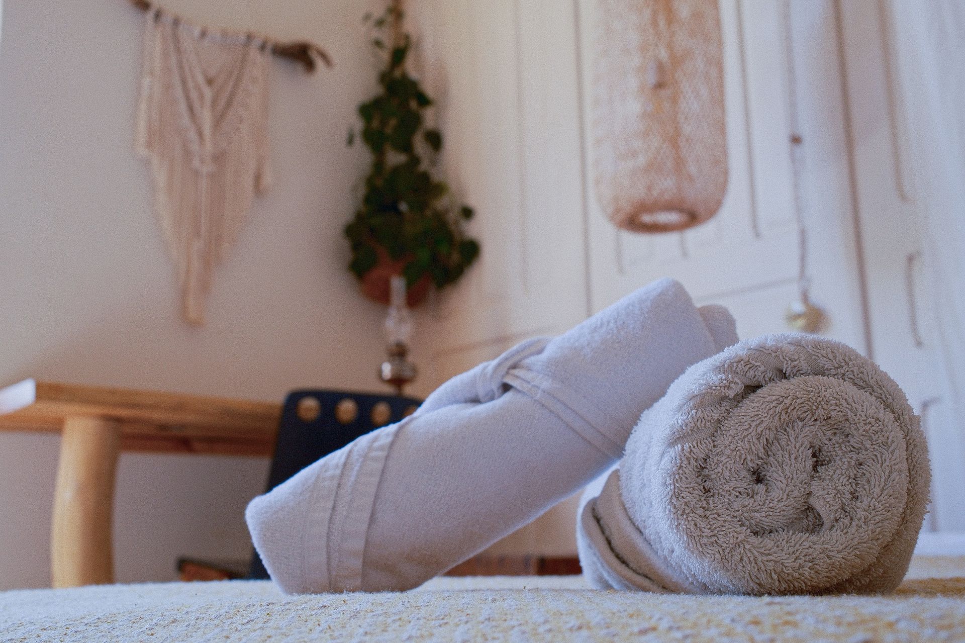 Two rolled-up beige towels rest on a surface in a cozy, softly lit room with a hanging plant and macrame wall decor.