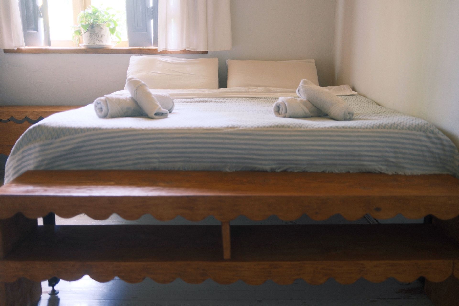 A made bed with two rolled towels and pillows, placed behind a wooden scalloped bench in a room with a window.