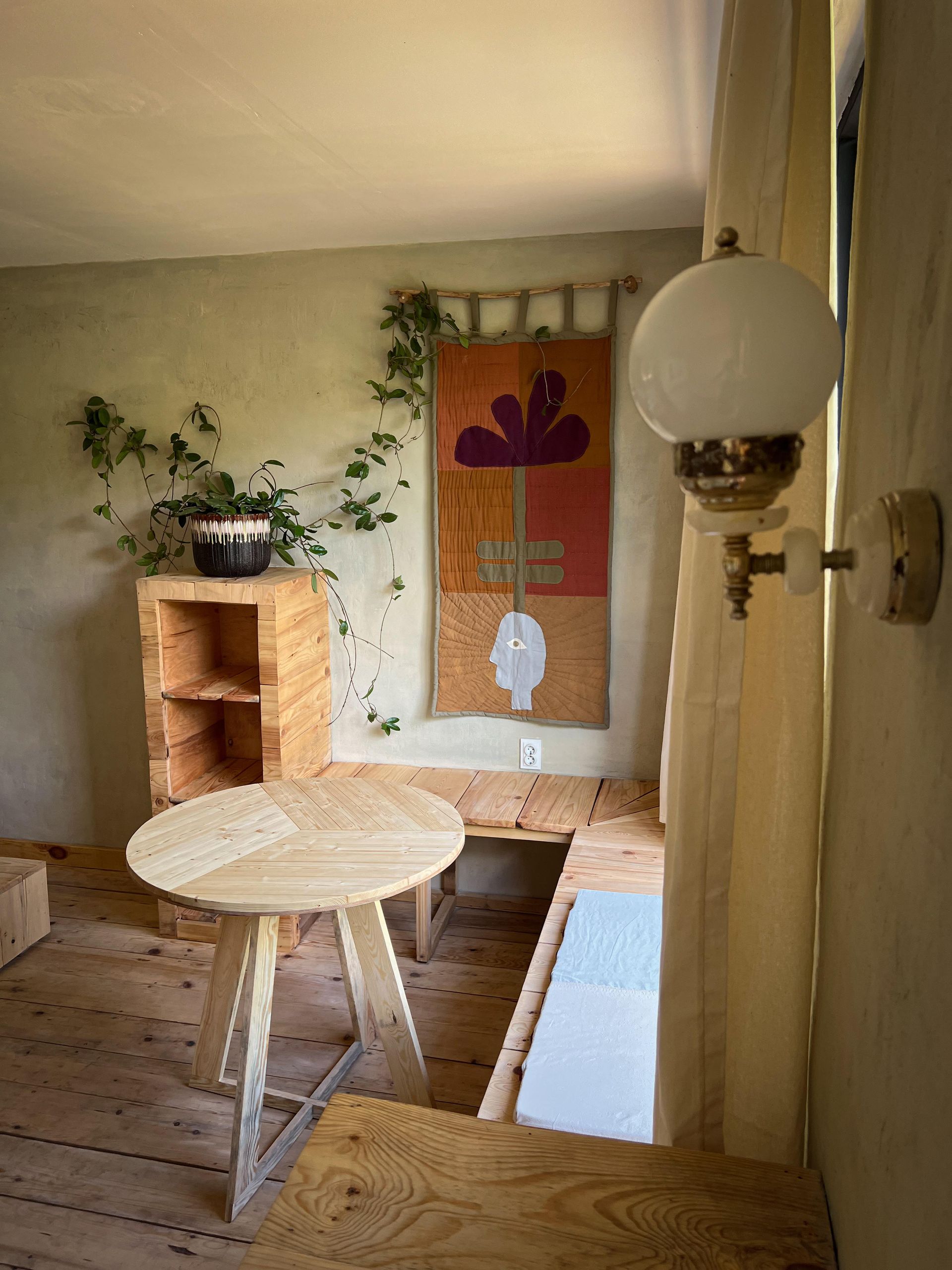 A small, rustic room features wooden furniture, a wall hanging of a purple flower, a potted plant, and a vintage lamp.