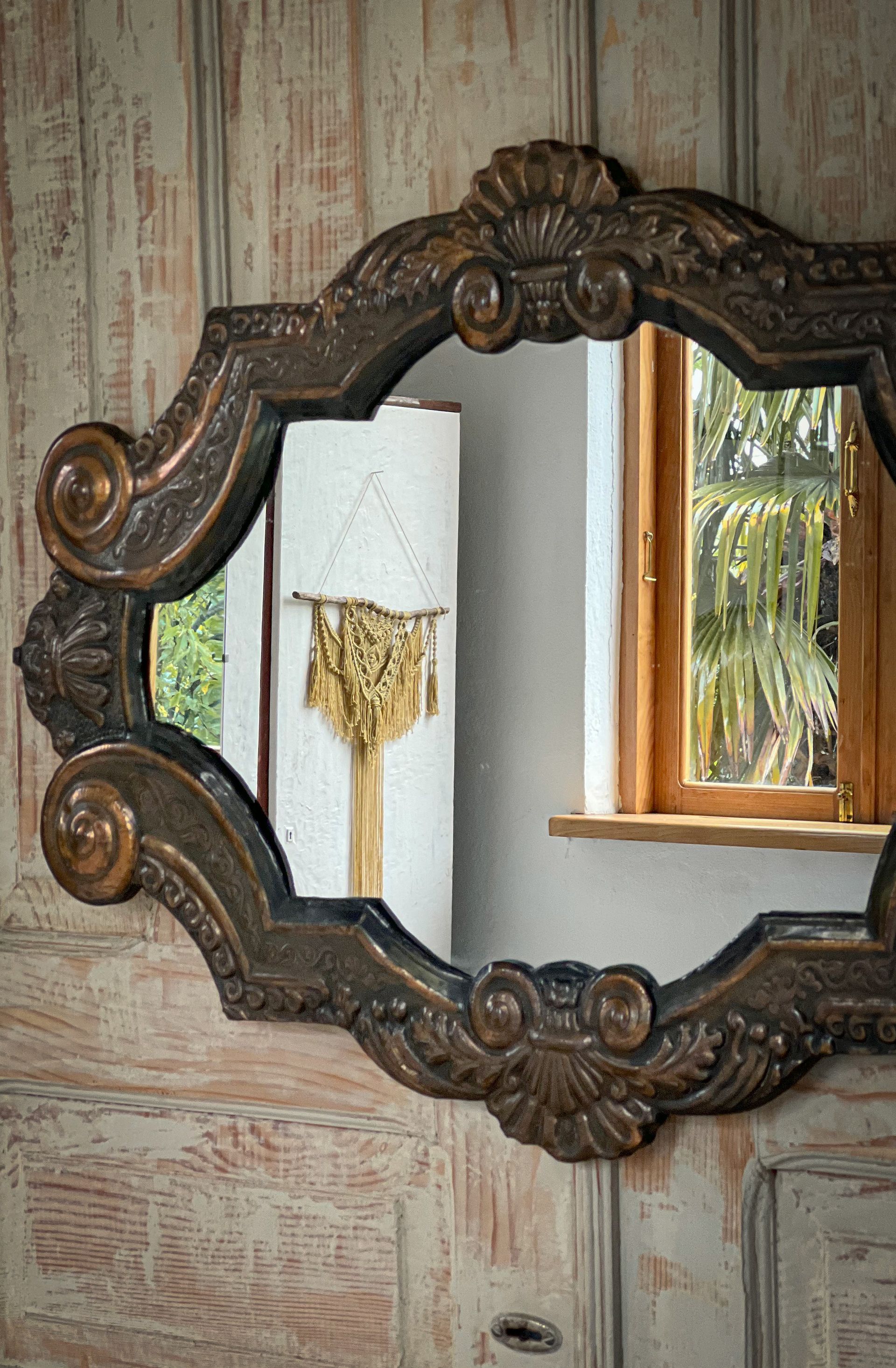 An ornate, dark-wood, scalloped-edge mirror hangs on a rustic wooden wall, reflecting a window and hanging macramé decor.