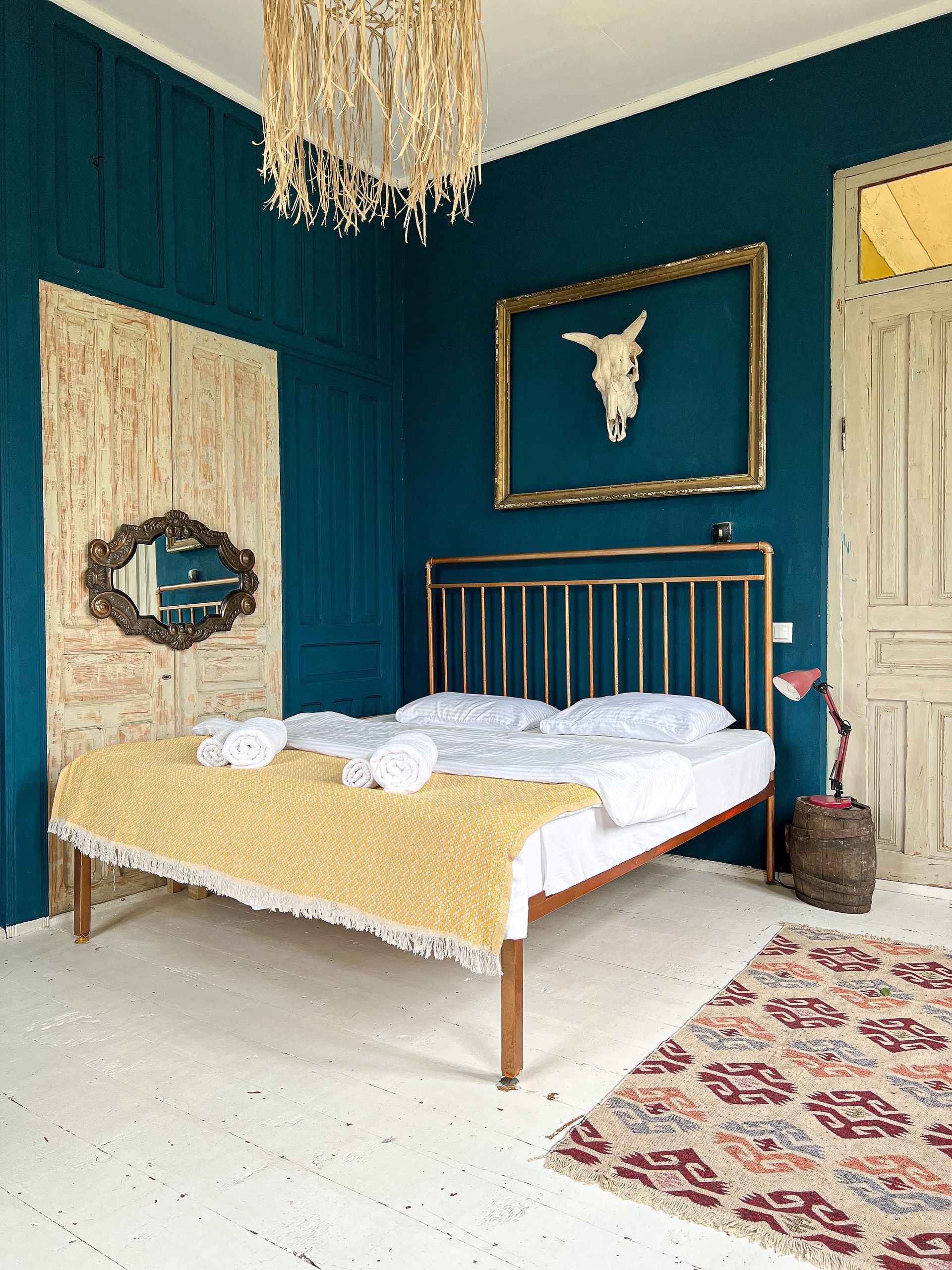 A bedroom with dark blue walls, a bed with a yellow throw blanket, a gold-framed skull, and a bohemian-style rug.
