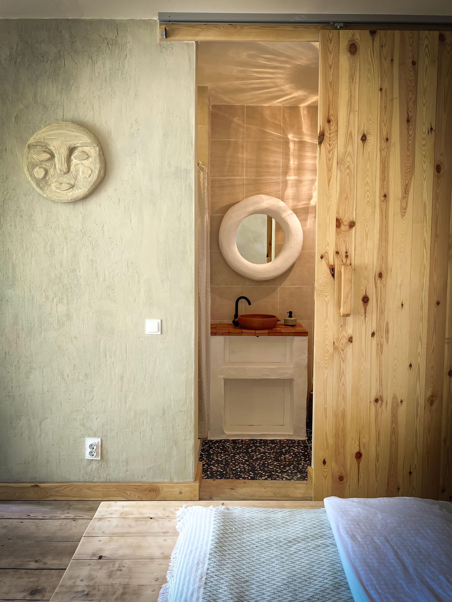 A bathroom seen through a wooden sliding door, featuring a round mirror, vanity, and a sun-faced wall art in the bedroom.