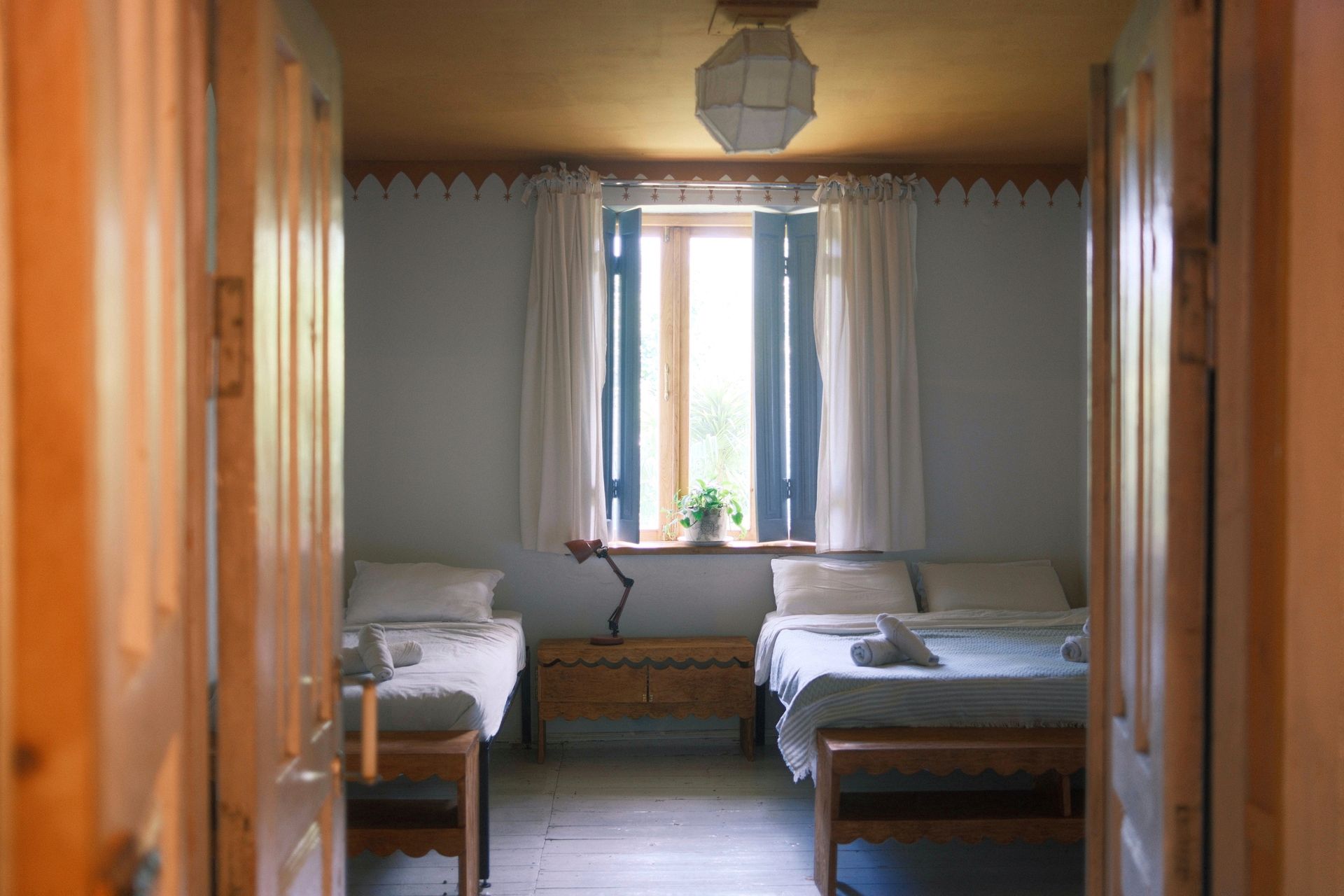 A bedroom view from a doorway, featuring two beds with white linens, a wooden dresser, and a window with white curtains.