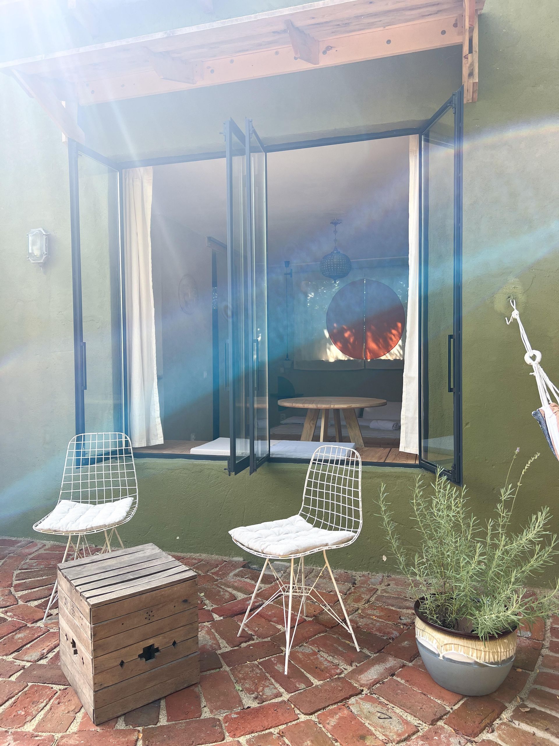 A brick patio with two white wire chairs, a wooden crate, and a potted plant in front of a glass door opening to a room.