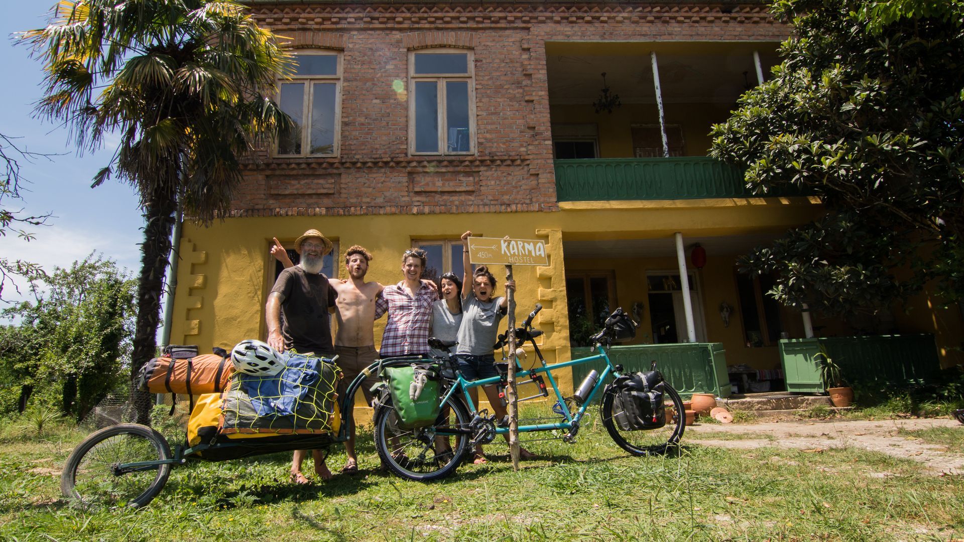 Five people pose in front of a yellow house with a tandem bicycle and a bicycle trailer loaded with gear.