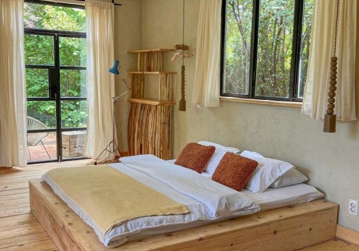 A minimalist bedroom with a wooden platform bed, white bedding, rust-colored pillows, and floor-to-ceiling windows.