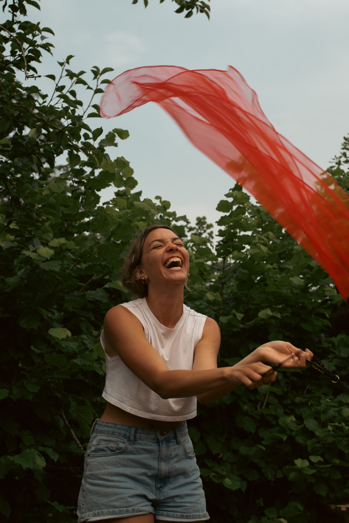 A person laughing outdoors while tossing a long, translucent red scarf into the air against a backdrop of green trees.