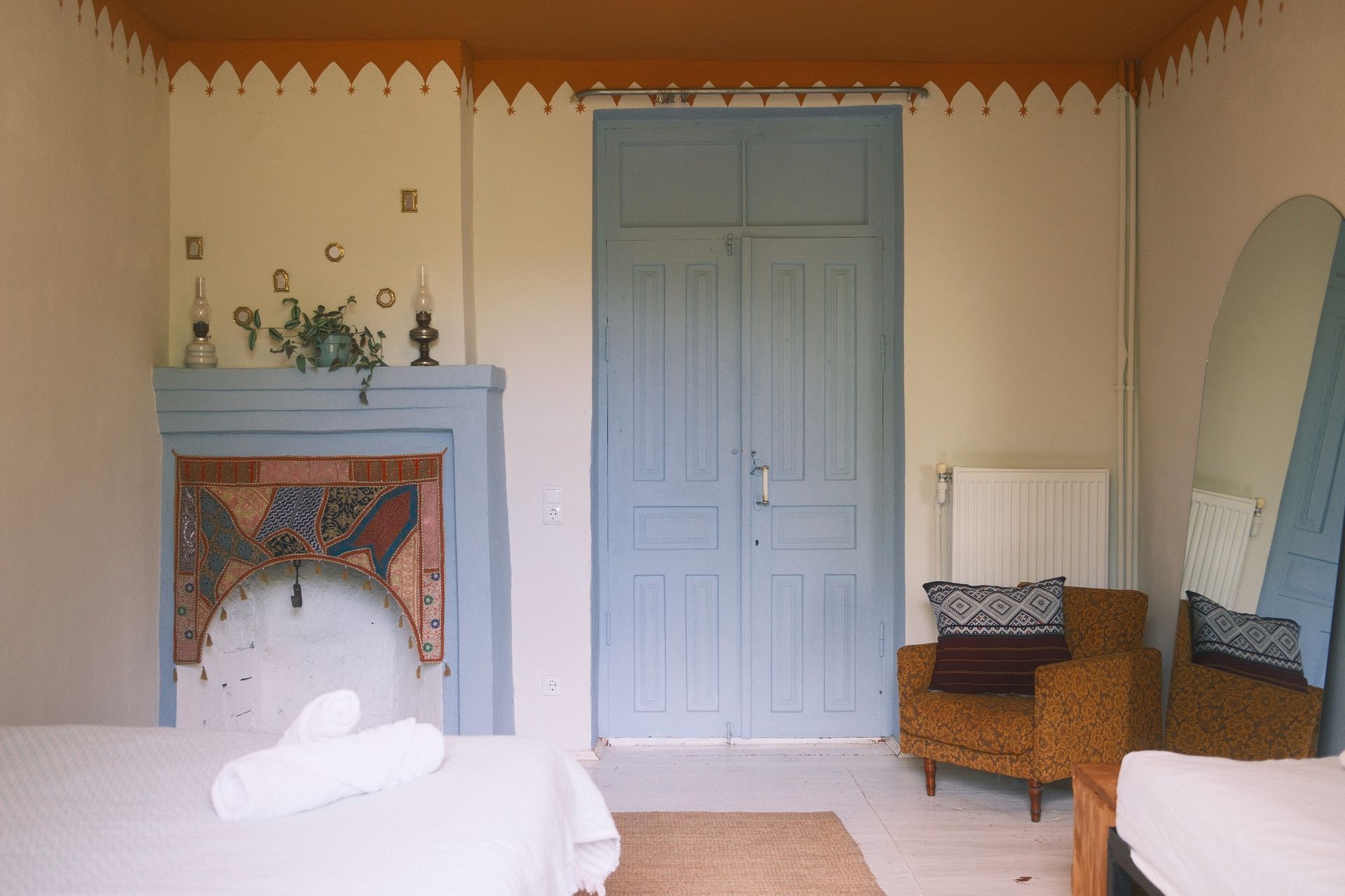 A bedroom with a fireplace, light blue double doors, an arched mirror, and an armchair with a patterned cushion.