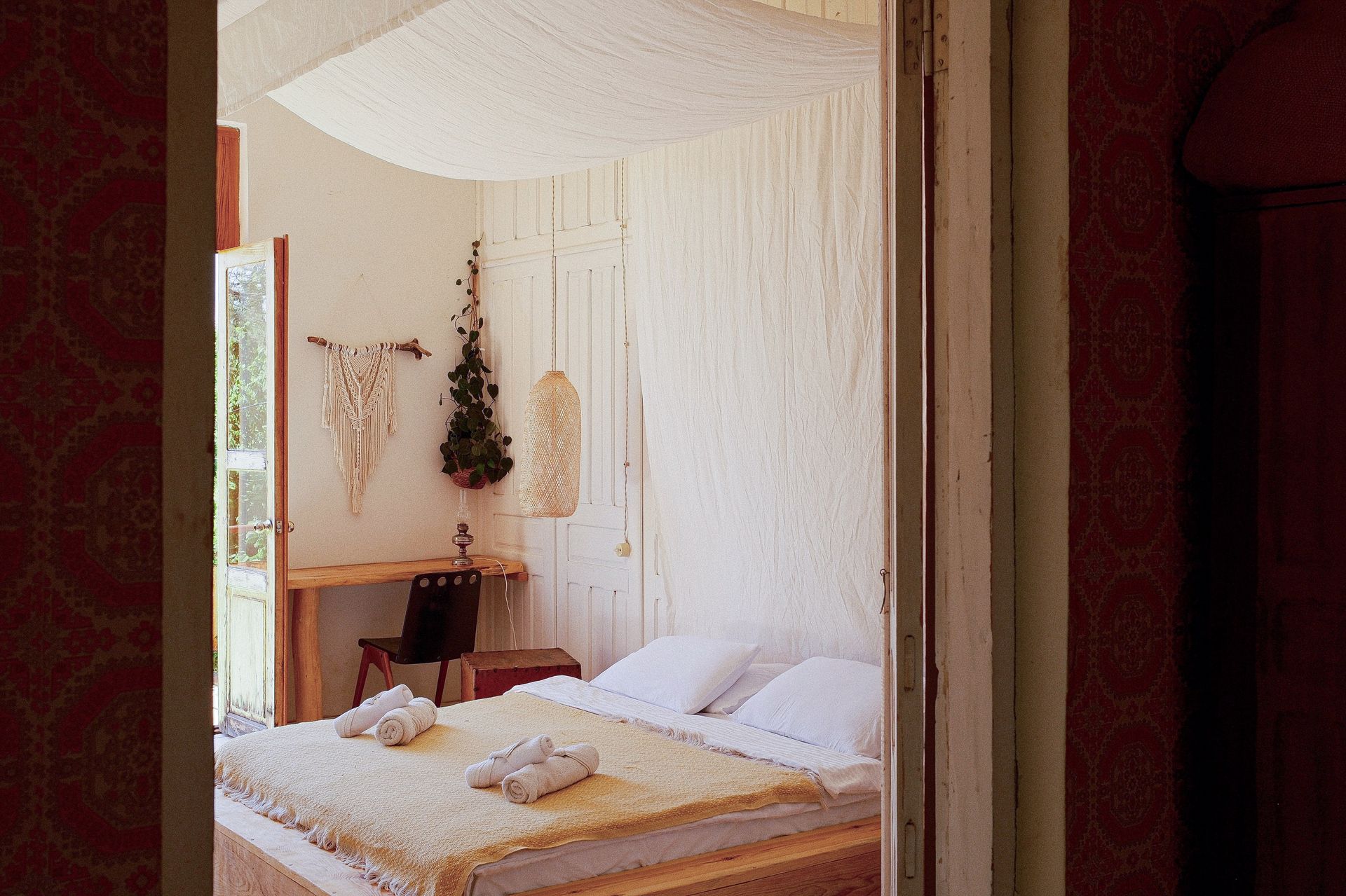 A bedroom with white walls and a canopy bed, seen through a doorway, with a desk, chair, and decorative macrame.