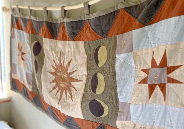 A patchwork wall hanging features sun, moon, and star motifs in shades of rust, sage green, and light blue.