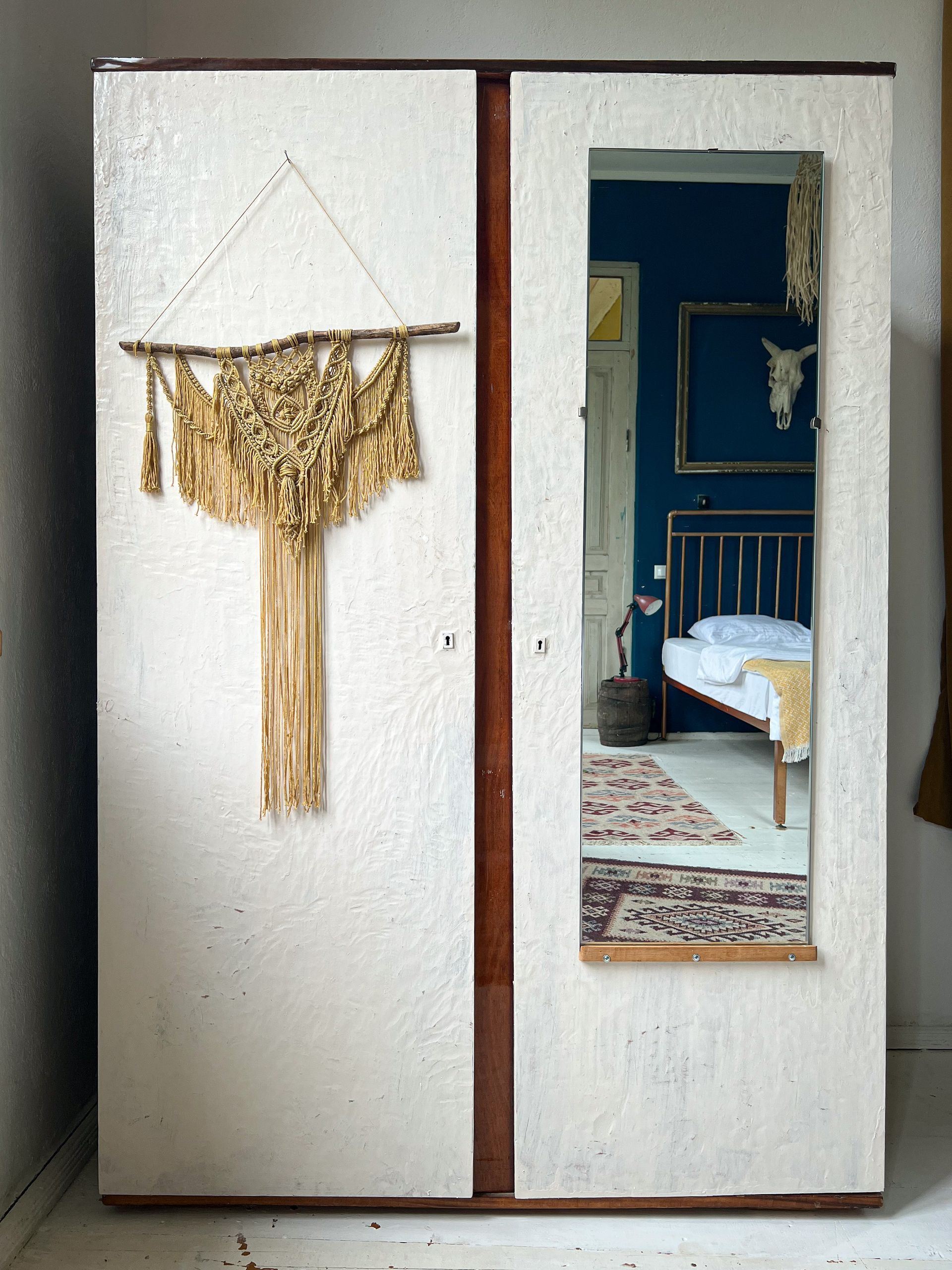 A two-door white wardrobe with a macramé wall hanging on the left and a mirror on the right reflecting a bedroom.