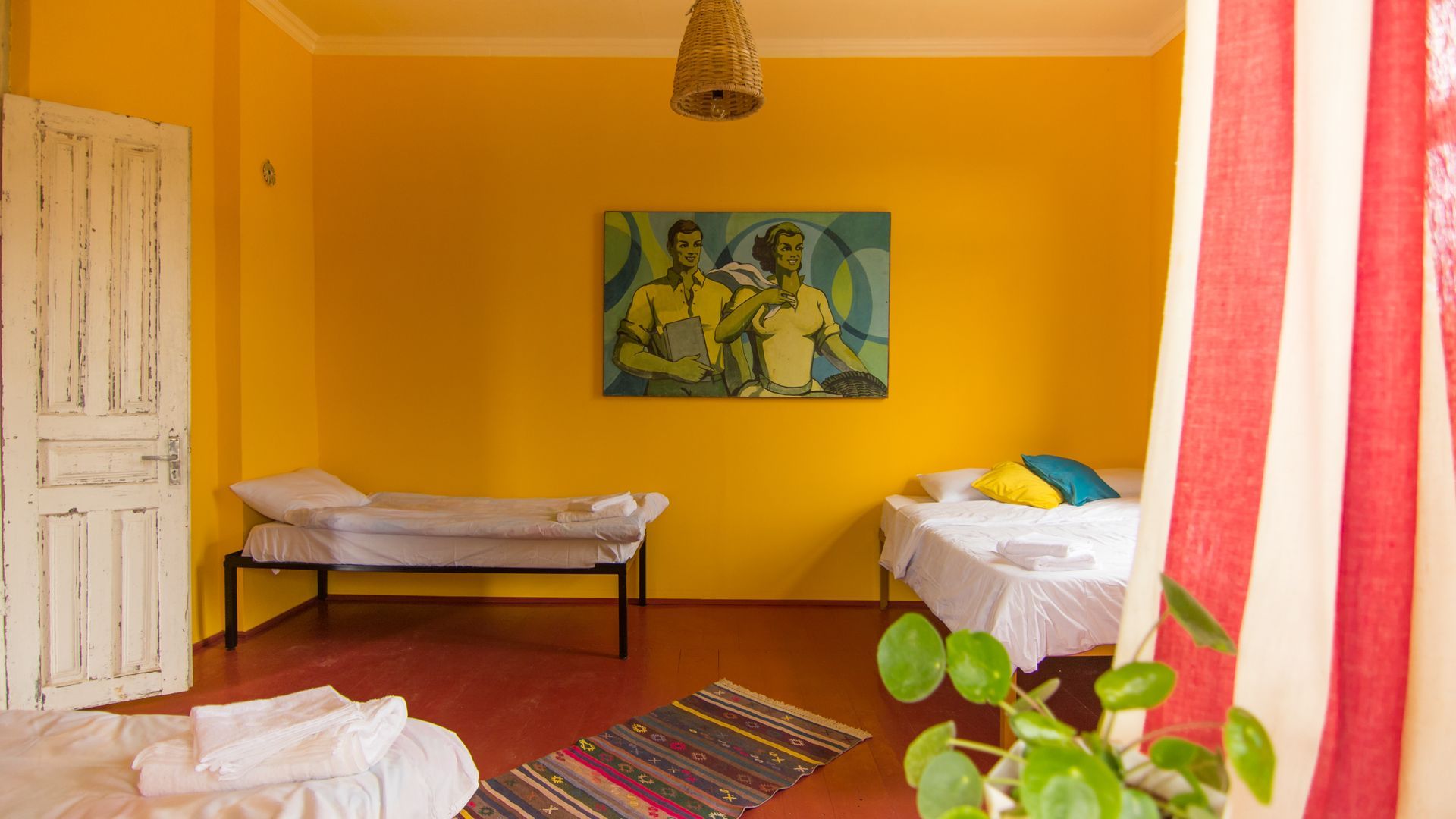 A bedroom with bright yellow walls, three beds, a patterned rug, and a painting featuring two figures on the wall.