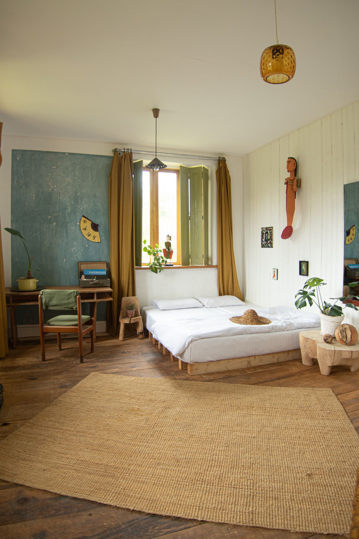 A rustic bedroom featuring a platform bed, wooden floors, a large jute rug, and a desk against a textured blue wall.