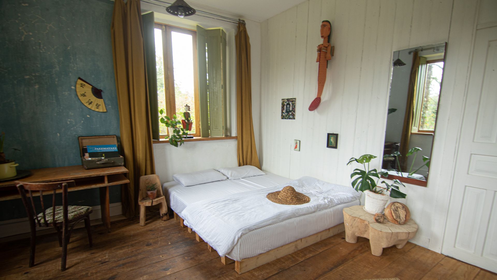 A cozy bedroom with a white bed, wooden furniture, a blue accent wall, a large mirror, and a window with mustard curtains.