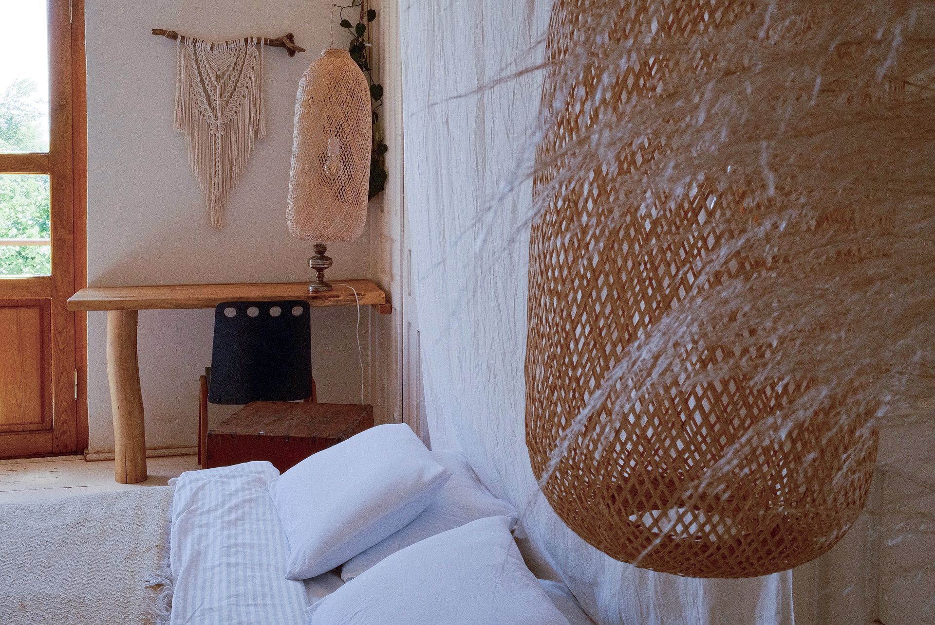 A cozy, sunlit room features a low bed with white linens, a woven pendant light, and a wooden desk with a macramé wall art.