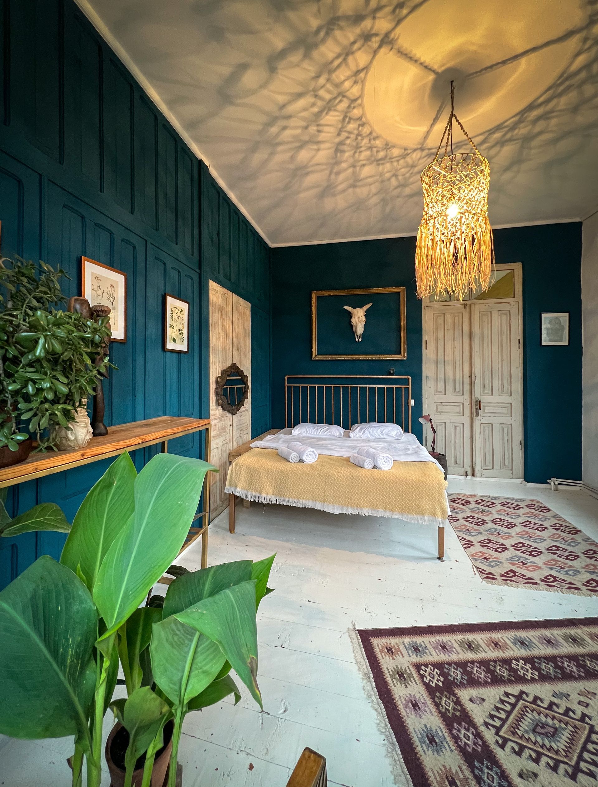 Bedroom with dark teal wood-paneled walls, a double bed, rustic light fixture, patterned rugs, and indoor plants.