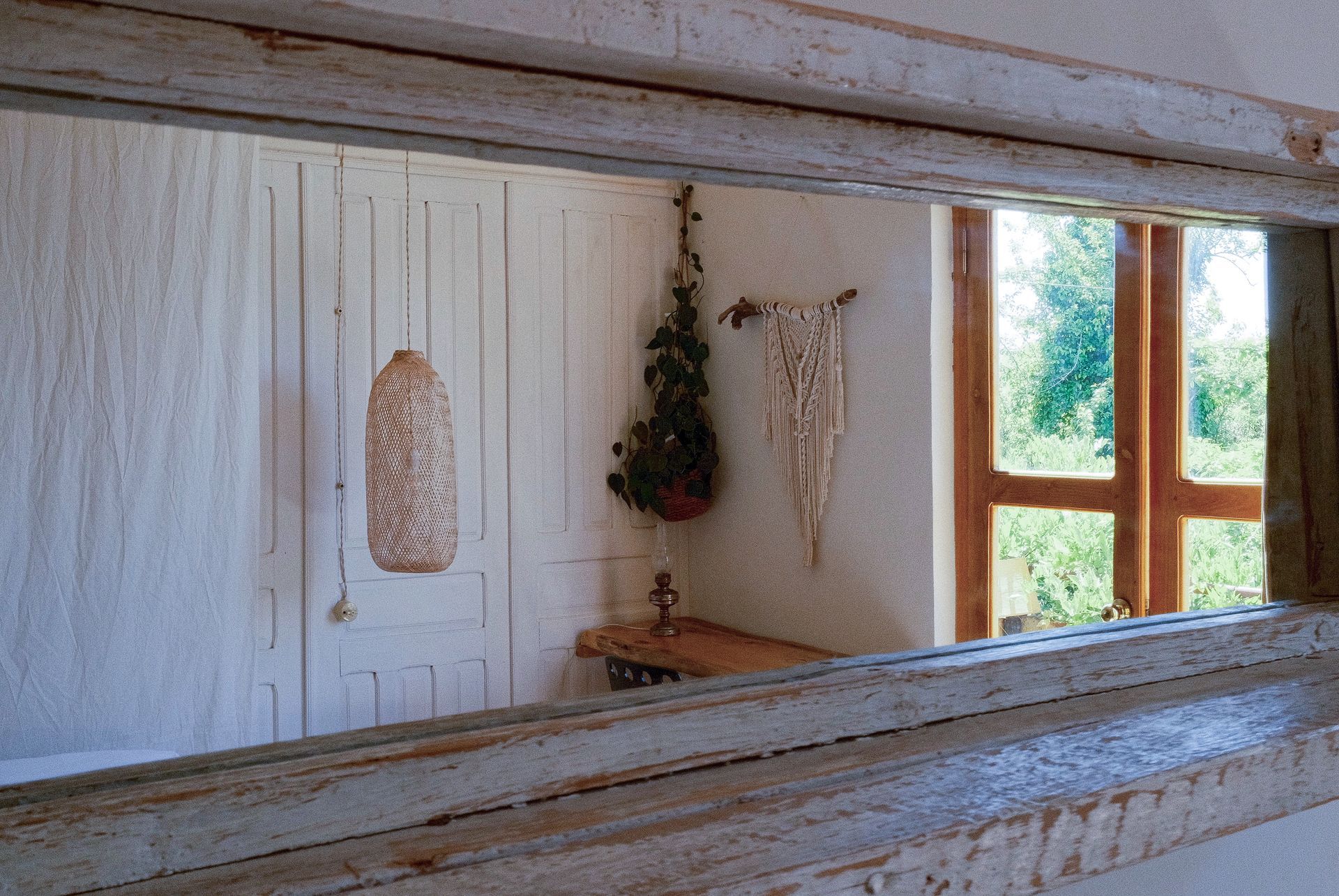 A wooden-framed mirror reflects a sunlit interior room featuring a hanging pendant light, wall plant, and macrame decor.