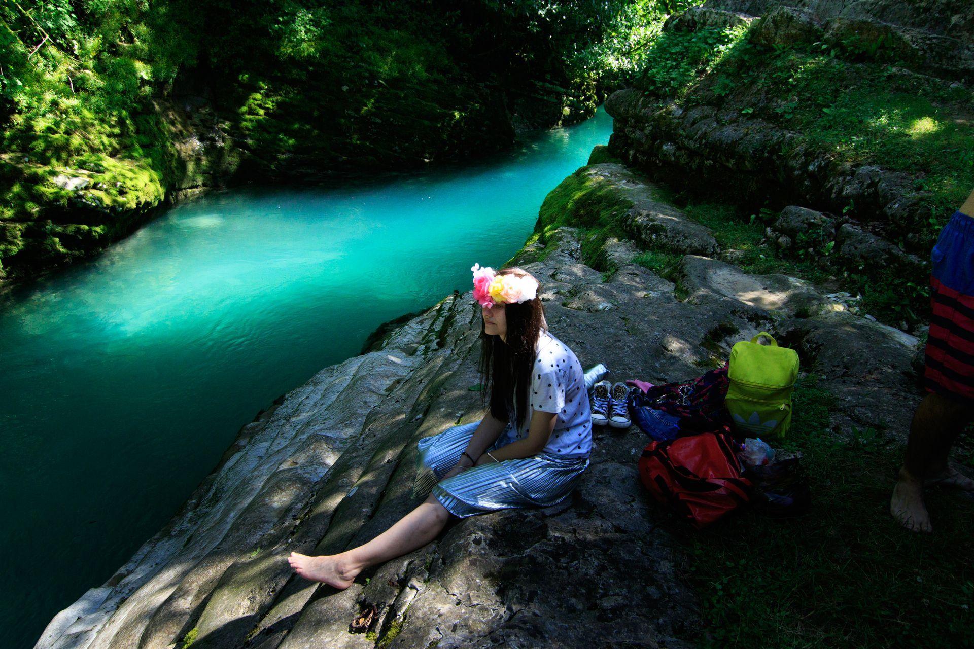 A person with a flower crown sits on a rocky ledge overlooking a vibrant, turquoise river surrounded by lush greenery.