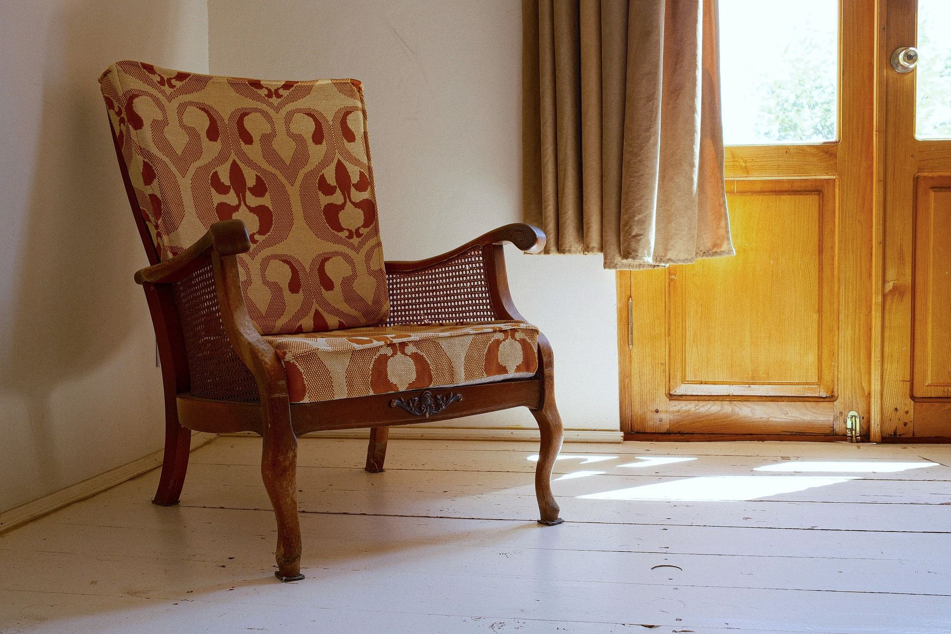 An upholstered armchair with a tan and maroon floral pattern stands on a white floor next to a window with tan curtains.