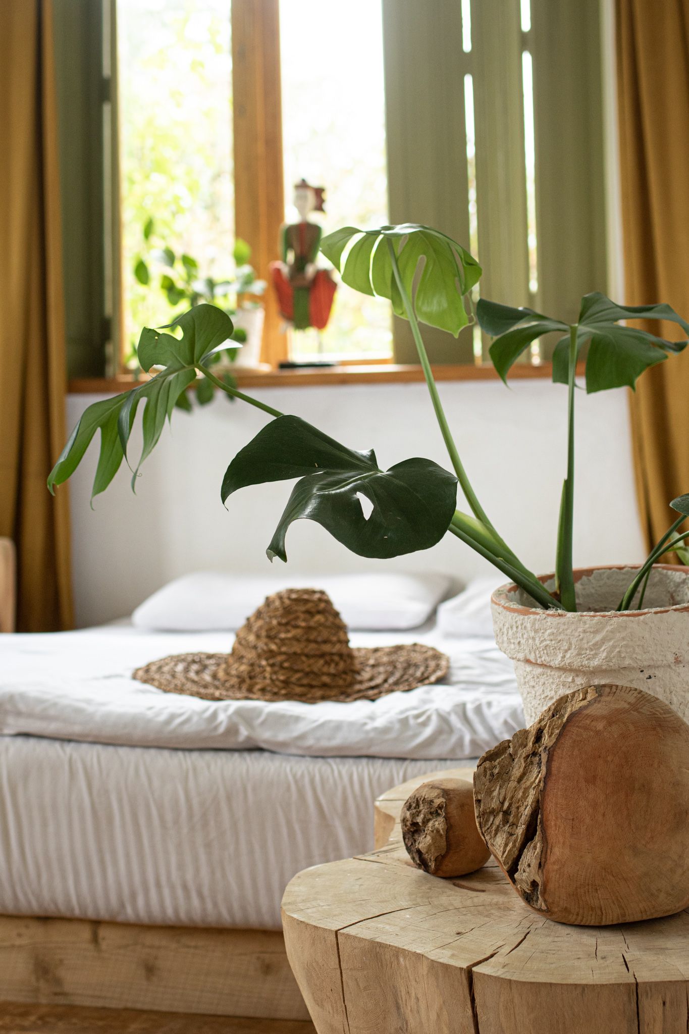 A sunlit bedroom features a bed with white linens, a straw hat, a potted Monstera plant, and a rustic wooden side table.