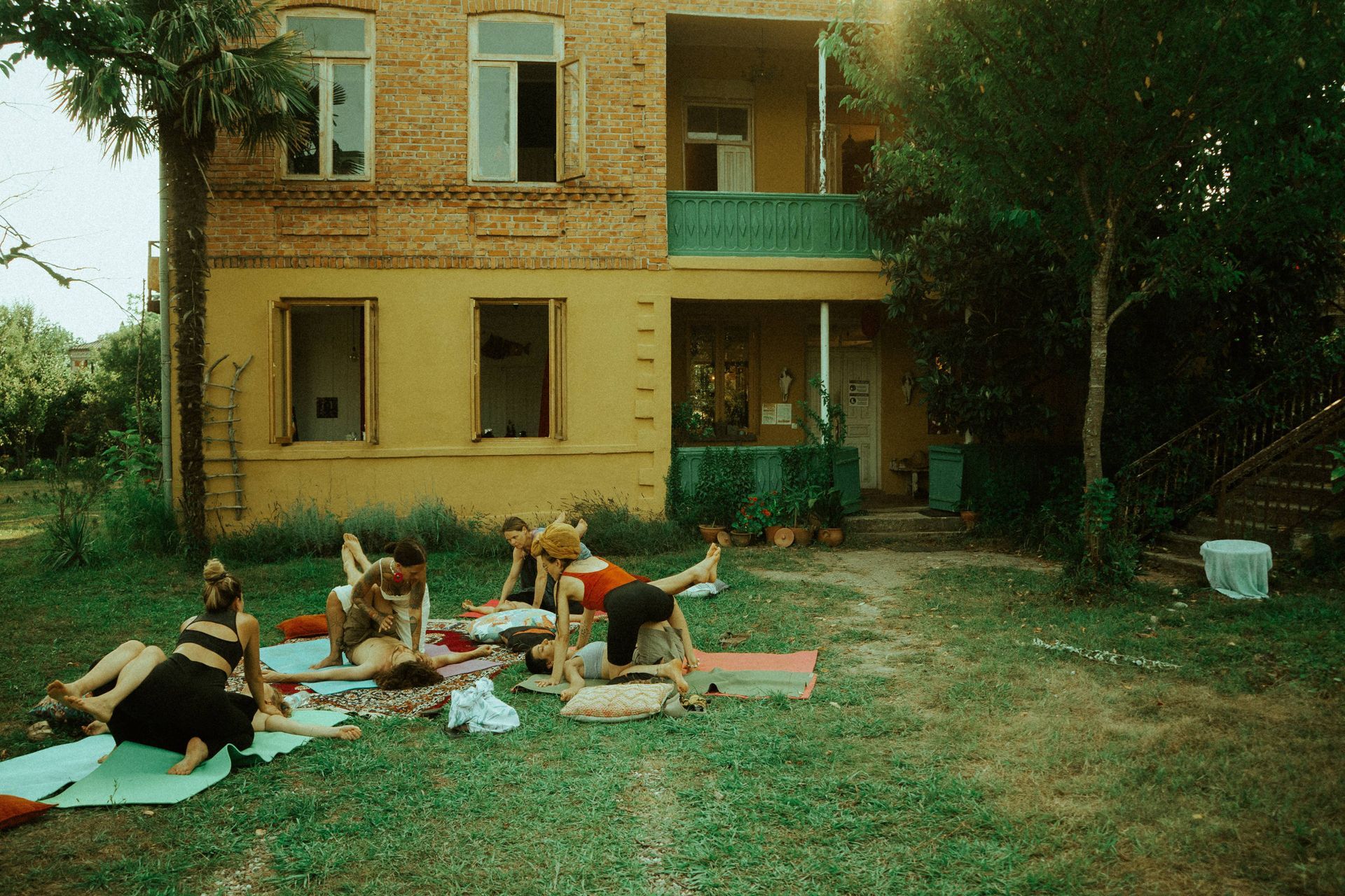 A group of people practices yoga on mats in a grassy yard in front of a yellow two-story house.
