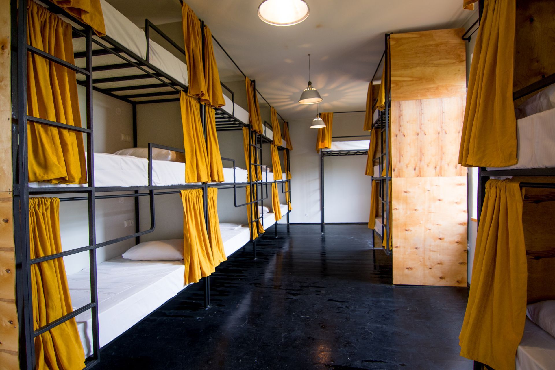 A hostel dormitory room featuring black metal bunk beds with white mattresses and yellow curtains on a dark floor.