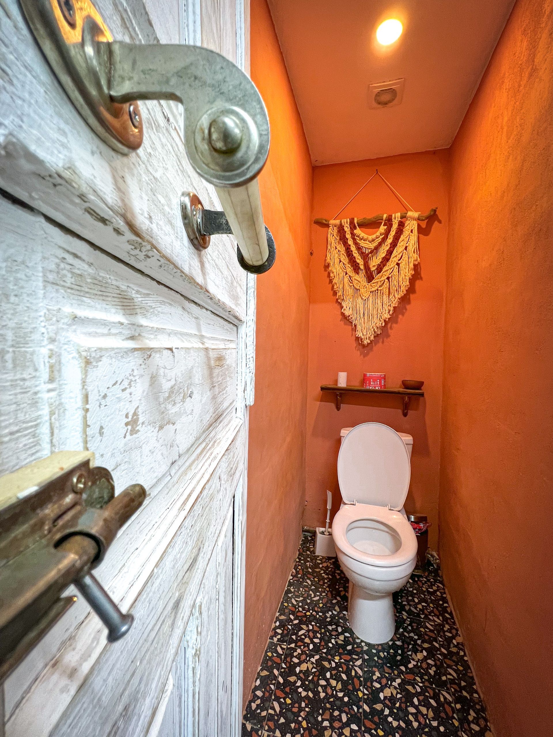 A view through a wooden door showing a narrow restroom with terracotta-colored walls, a toilet, and macramé decor.