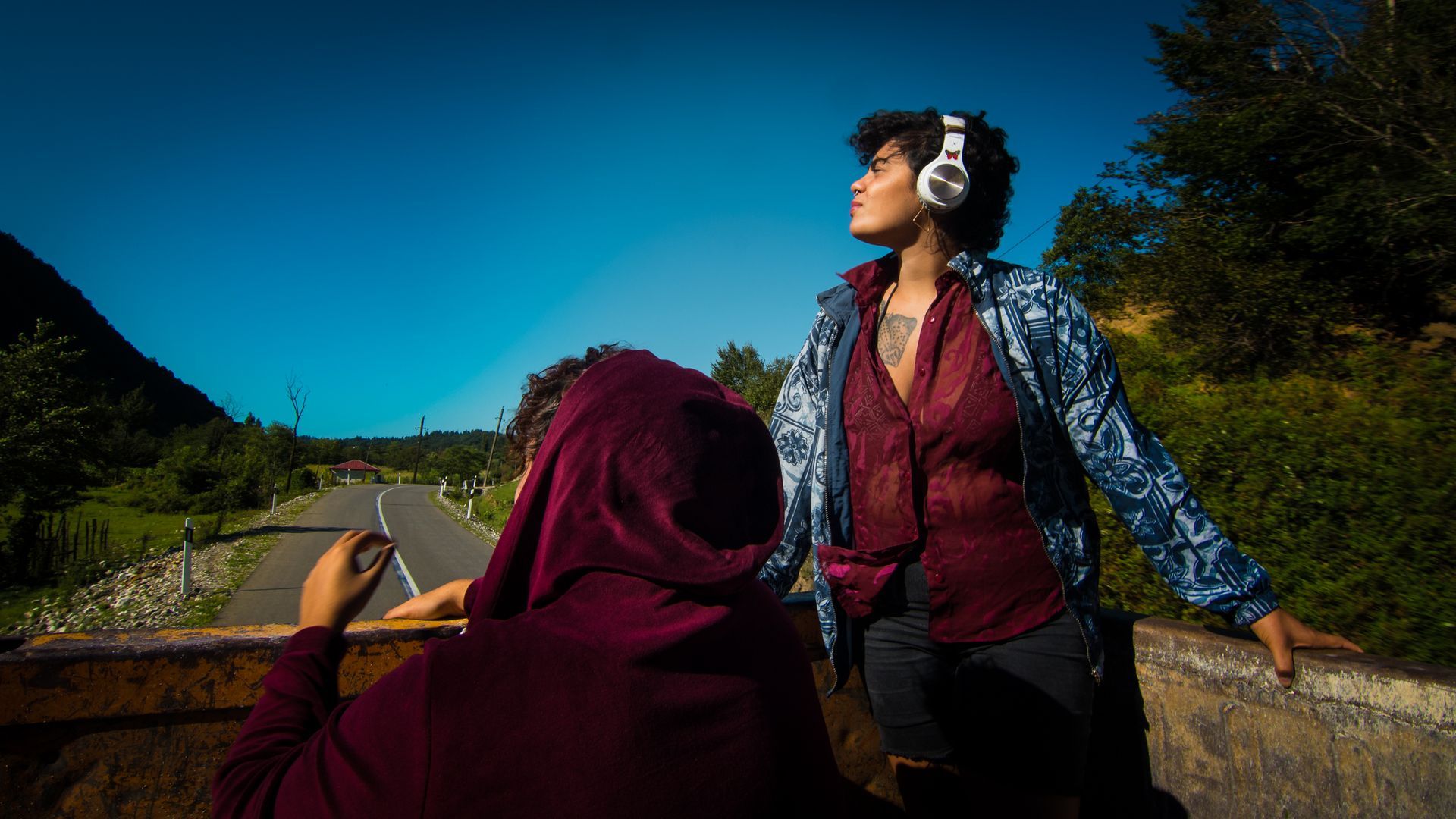 Two people ride in the back of a vehicle on a rural road, one wearing headphones and looking toward the sky.