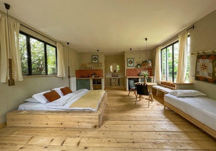 A rustic, open-plan bedroom with a low wooden platform bed, a single bed, and a small dining area on a wooden floor.