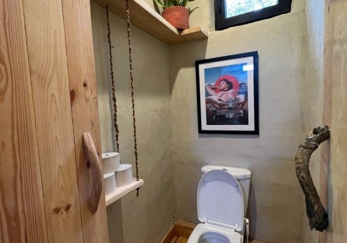 A small bathroom featuring a toilet, a hanging shelf for toilet paper, and a framed art print on a textured beige wall.