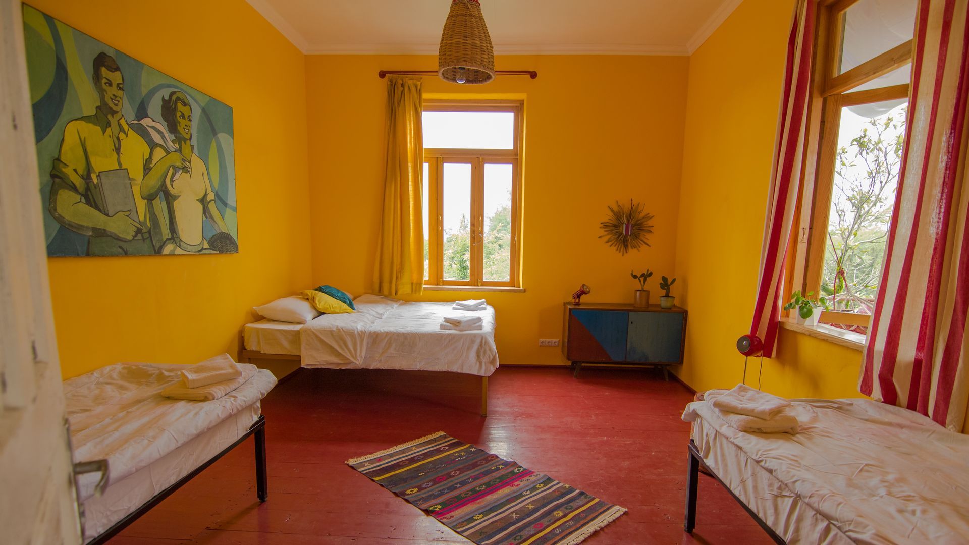 A room with yellow walls, a red floor, three beds, a central rug, a large painting, and striped curtains by a window.