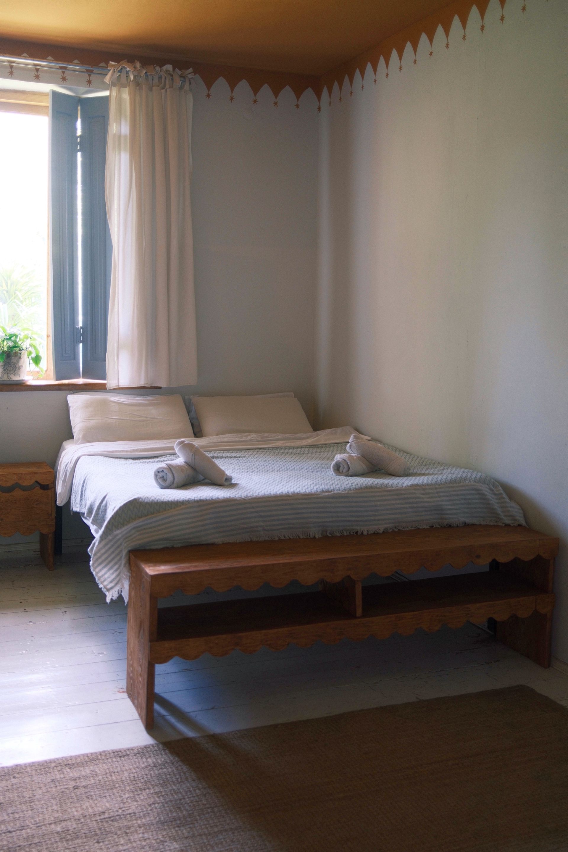 A neatly made bed with white bedding and two towels, in a room with light walls and a wooden bench at the foot.