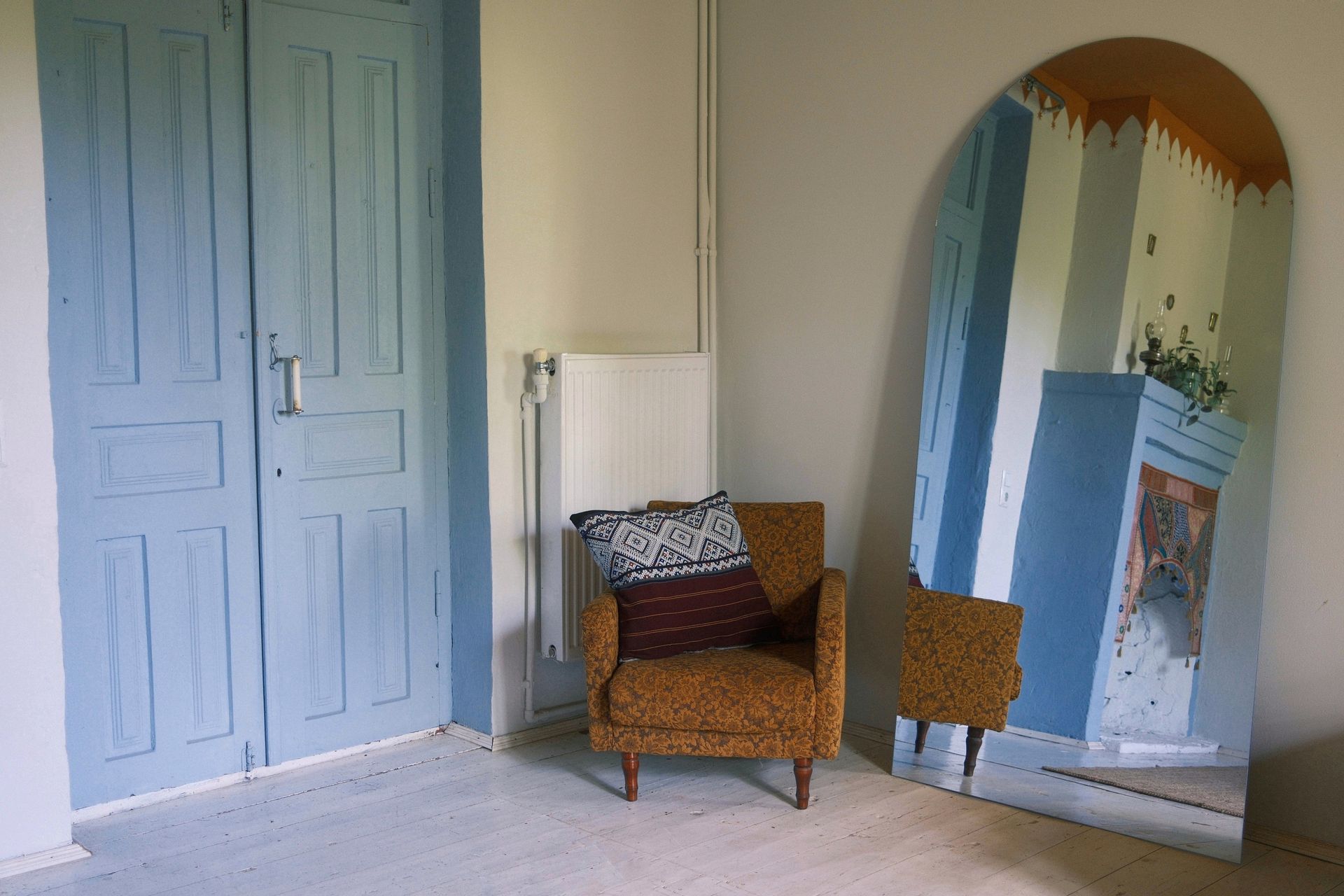 A mustard-colored armchair with a patterned cushion sits on a white floor next to a blue door and an arched floor mirror.