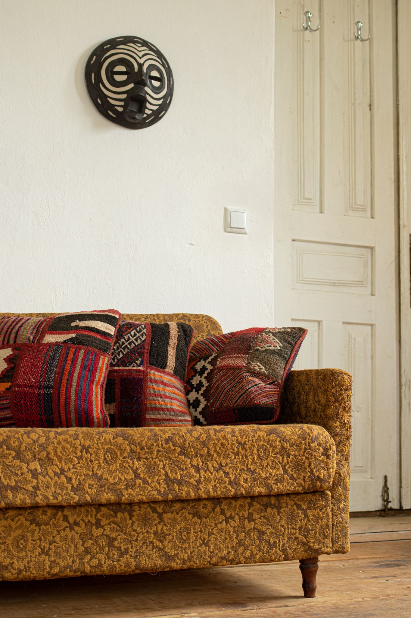 A textured, mustard-yellow sofa with patterned throw pillows sits beneath a tribal-style mask hanging on a white wall.