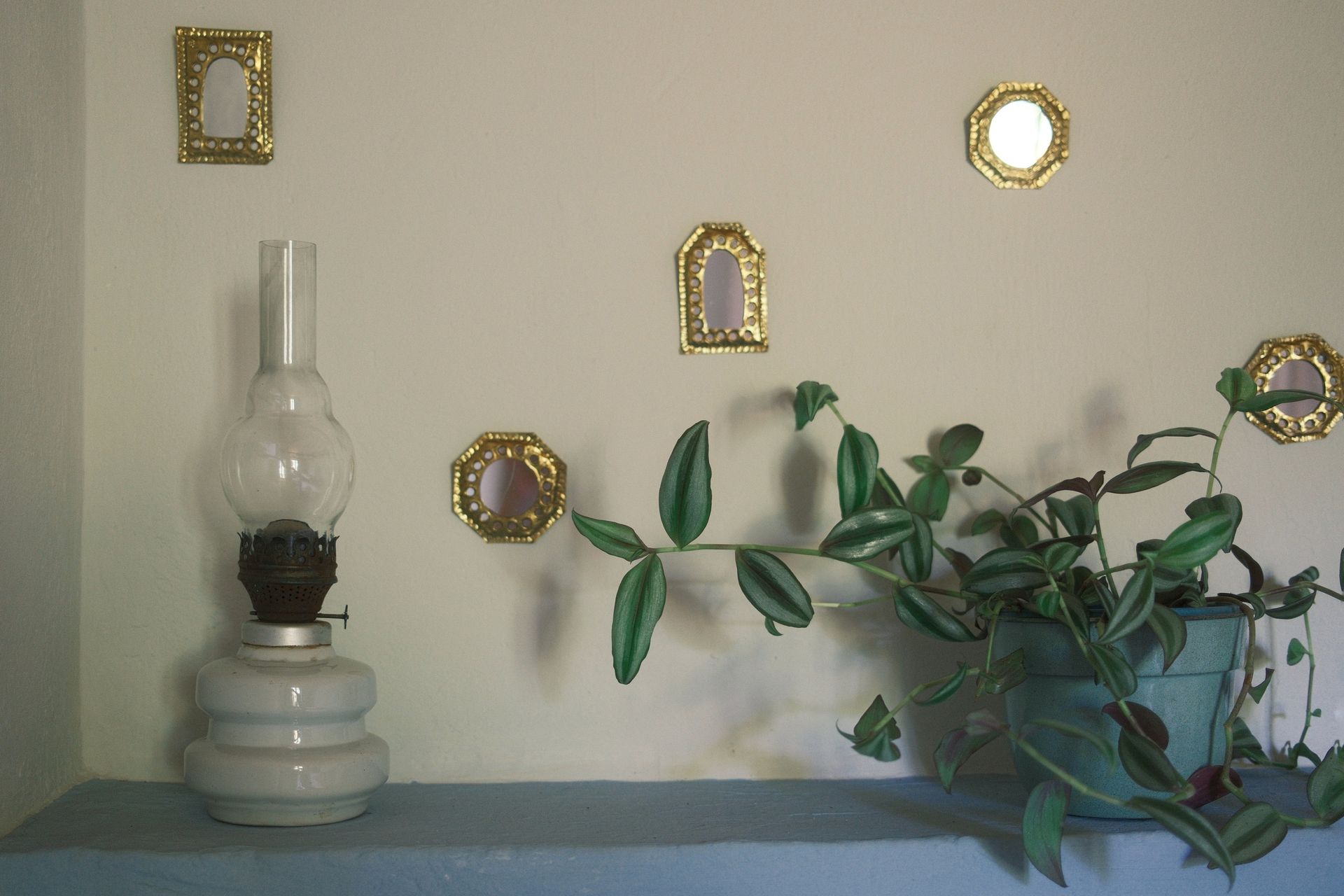 A vintage kerosene lamp and a trailing houseplant sit on a blue shelf against a cream wall decorated with gold mirrors.