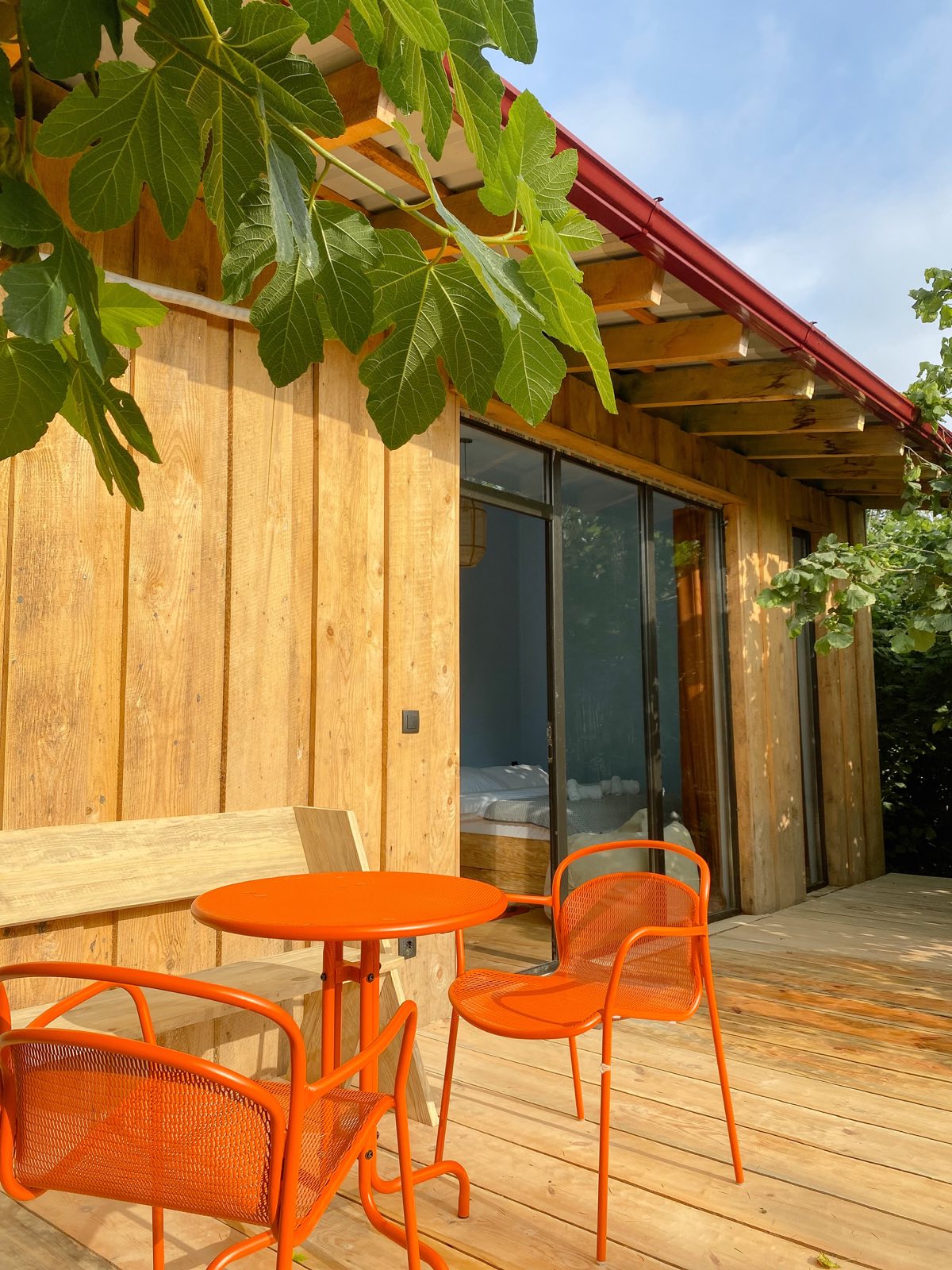 A wooden cabin with a private terrace, an orange bistro table and chairs under a hazelnut tree.